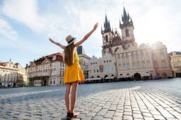 Young female tourist dressed in yellow standing with raised hands on the old town square of Prague. Enjoying great vacation in Czech republic